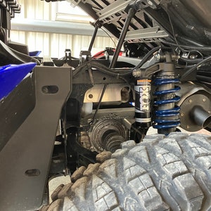 Puede incluir: Primer plano de la suspensión trasera de un vehículo todoterreno azul. La suspensión cuenta con un amortiguador negro FOX Performance Series con un muelle helicoidal azul. El vehículo está estacionado en un garaje.