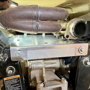 Puede incluir: Primer plano de una placa de deslizamiento de metal plateado con dos tornillos en el lado derecho. La placa de deslizamiento está montada en un marco de metal negro con un tubo de escape marrón encima.