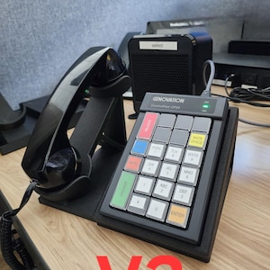 May include: A black Genovation ControlPad CP24 keypad with a handset resting on a stand. The keypad features a grid of square keys with alphanumeric labels, including coloured keys labelled Release, Mute, Answer, Back Space, and Enter.