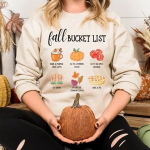 Op de afbeelding: Een beige sweatshirt met de tekst "Fall Bucket List" en illustraties van herfstactiviteiten. Het sweatshirt heeft een pumpkin spice latte, pompoenen, appels, wandelschoenen, herfstbladeren en een taart. Een persoon houdt een decoratieve pompoen vast.