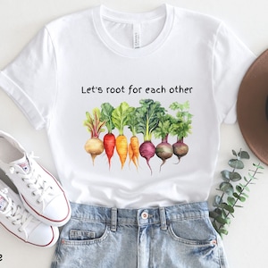 Op de afbeelding: Wit T-shirt met een aquarel-illustratie van groenten en de tekst "Let's root for each other".
