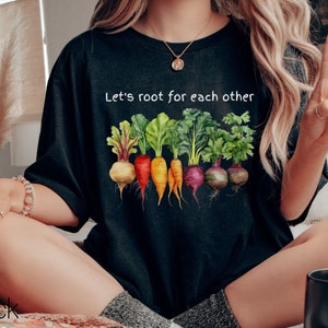 Op de afbeelding: Een zwart T-shirt met de tekst "Let's root for each other" en een afbeelding van kleurrijke wortelgroenten.