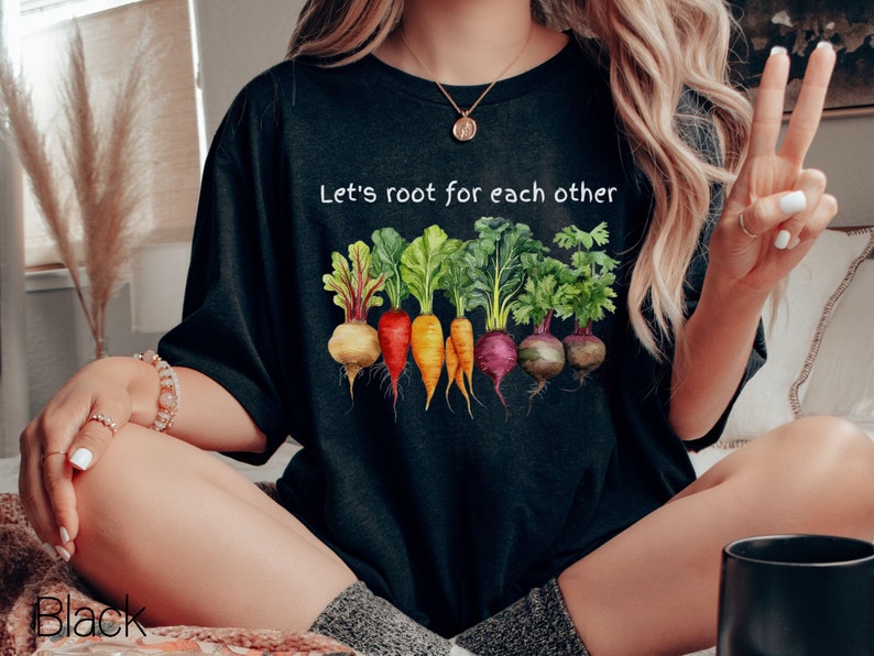 Op de afbeelding: Een zwart T-shirt met de tekst "Let's root for each other" en een afbeelding van kleurrijke wortelgroenten.