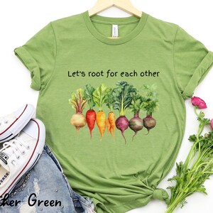 Op de afbeelding: Een groen T-shirt met de tekst "Let's root for each other" en een aquarel-illustratie van kleurrijke wortelgroenten.
