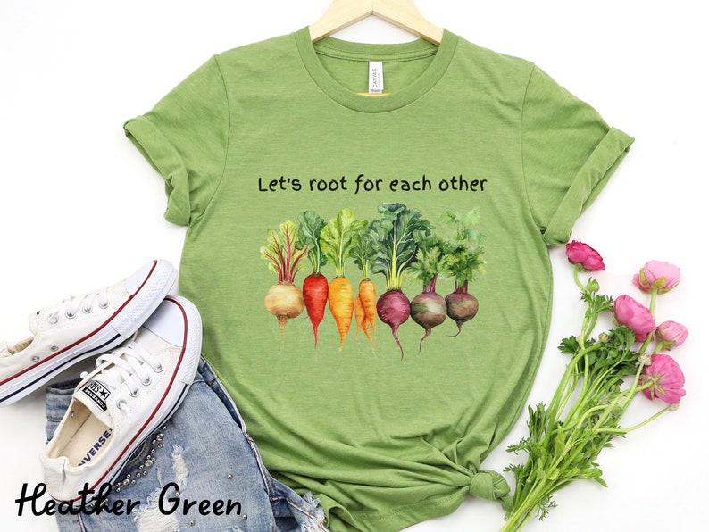 Op de afbeelding: Een groen T-shirt met de tekst "Let's root for each other" en een aquarel-illustratie van kleurrijke wortelgroenten.
