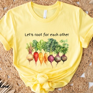 Op de afbeelding: Geel T-shirt met de tekst "Let's root for each other" en een afbeelding van kleurrijke wortelgroenten.