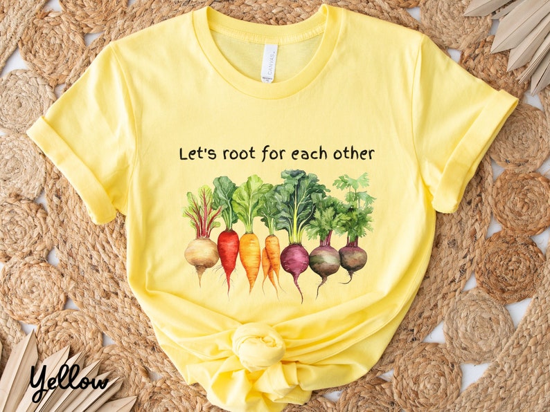 Op de afbeelding: Geel T-shirt met de tekst "Let's root for each other" en een afbeelding van kleurrijke wortelgroenten.