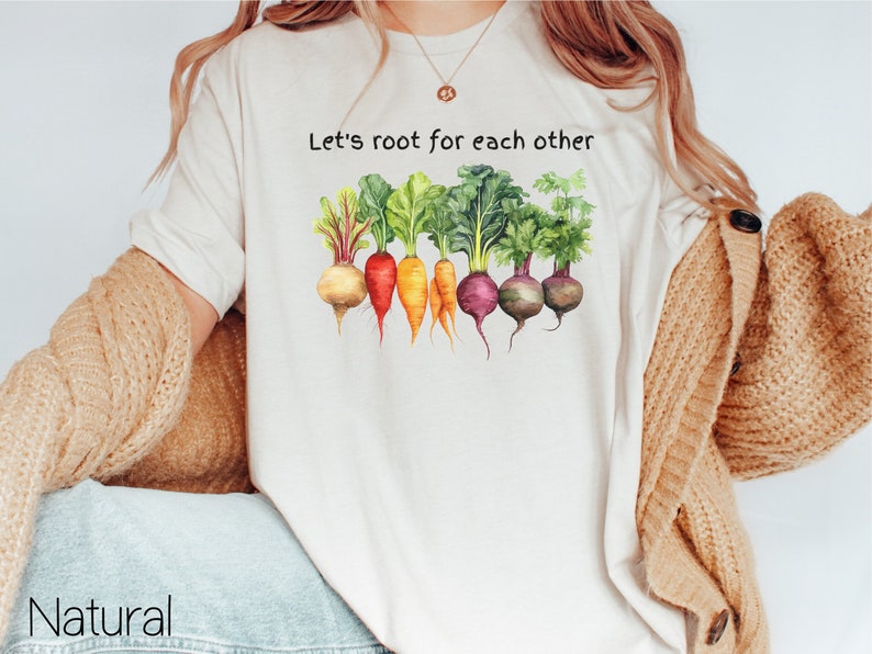 Op de afbeelding: Wit T-shirt met de tekst "Let's root for each other" en een aquarel-illustratie van een rij wortels en bieten.