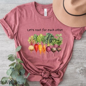 Op de afbeelding: Een heide-paarse t-shirt met de tekst "Let's root for each other" en een afbeelding van kleurrijke wortelgroenten.