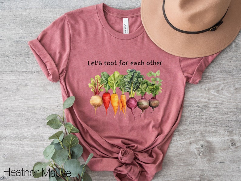 Op de afbeelding: Een heide-paarse t-shirt met de tekst "Let's root for each other" en een afbeelding van kleurrijke wortelgroenten.