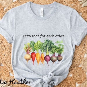 Op de afbeelding: Grijs T-shirt met de tekst "Let's root for each other" en een aquarel-illustratie van een rij kleurrijke wortelgroenten.