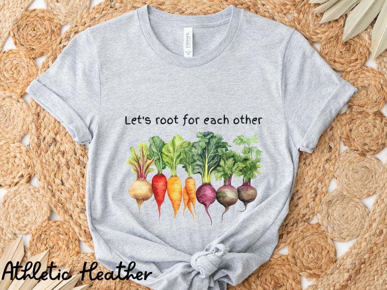 Op de afbeelding: Grijs T-shirt met de tekst "Let's root for each other" en een aquarel-illustratie van een rij kleurrijke wortelgroenten.