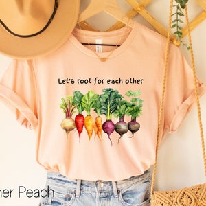 Op de afbeelding: Een perzikkleurig T-shirt met de tekst "Let's root for each other" en een afbeelding van kleurrijke wortelgroenten.