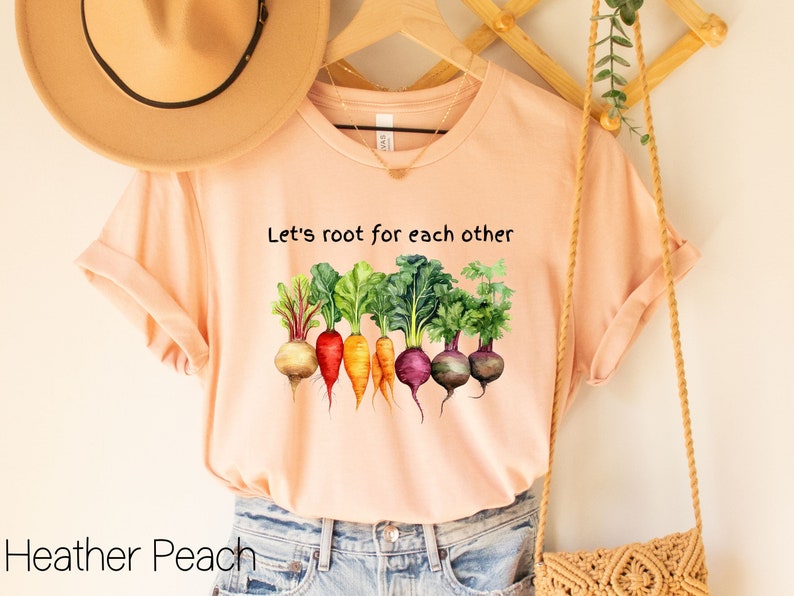 Op de afbeelding: Een perzikkleurig T-shirt met de tekst "Let's root for each other" en een afbeelding van kleurrijke wortelgroenten.
