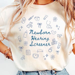 May include: Ivory-colored t-shirt with the text "Newborn Hearing Screener" in blue script, surrounded by blue line-art illustrations of baby-related items like a bottle, stethoscope, and baby feet.