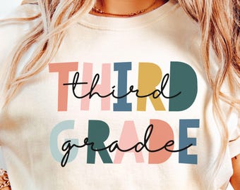 May include: Ivory-colored t-shirt with the words "THIRD GRADE" in colorful block letters and cursive script. The text is in shades of pink, blue, yellow, and green. A casual, comfortable top for students or teachers.