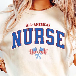 Op de afbeelding: Ivoor kleurige sweatshirt met de tekst "ALL-AMERICAN NURSE" in rood en blauw. Twee Amerikaanse vlaggen worden onder de tekst weergegeven, vastgebonden met een blauw lint. Het woord "IVORY" staat rechtsonder.