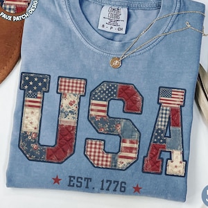 May include: A blue Comfort Colors t-shirt with "USA" in a faux patchwork design featuring the American flag and floral patterns. The shirt includes "EST. 1776" and is labeled "Blue Jean". A gold necklace rests on the shirt.