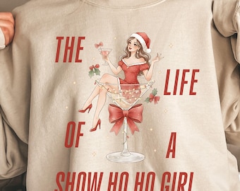 Op de afbeelding: Zandkleurige sweatshirt met een afbeelding van een vrouw met een kerstmuts in een martini glas, die een cocktail vasthoudt. De tekst luidt "THE LIFE OF A SHOW HO HO GIRL" in het rood. Een grote rode strik siert het glas.