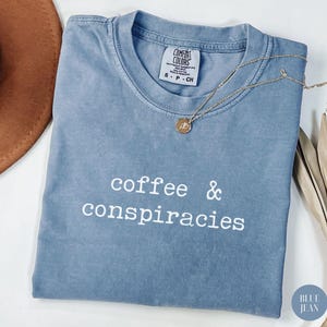 May include: A blue jean colored t-shirt with the words "coffee & conspiracies" printed in white. A gold necklace with a pendant rests on the shirt. The shirt has a Comfort Colors tag.