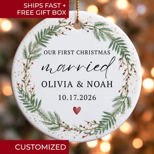 May include: A round white ceramic ornament featuring a green pine wreath with red berries and gold speckles. The text reads OUR FIRST CHRISTMAS married OLIVIA & NOAH 10.17.2026 with a small red heart below. A gold string is attached to the top.