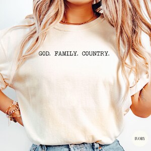 May include: Ivory-colored t-shirt with the text "GOD. FAMILY. COUNTRY." in a simple, black font. The shirt is a neutral tone, suitable for various styles. The image shows a close-up of the shirt.