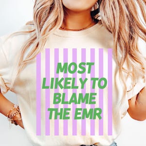 Op de afbeelding: Cr&egrave;mekleurig T-shirt met een verticaal gestreept ontwerp in lichtpaars. Het shirt heeft de tekst "MOST LIKELY TO BLAME THE EMR" in groene blokletters. Het shirt is gemaakt van een zacht materiaal.