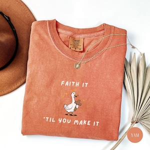 May include: A coral-colored Comfort Colors t-shirt with the phrase "Faith It 'Til You Make It" and a cartoon goose holding a cross. A gold necklace and a brown hat are also visible.