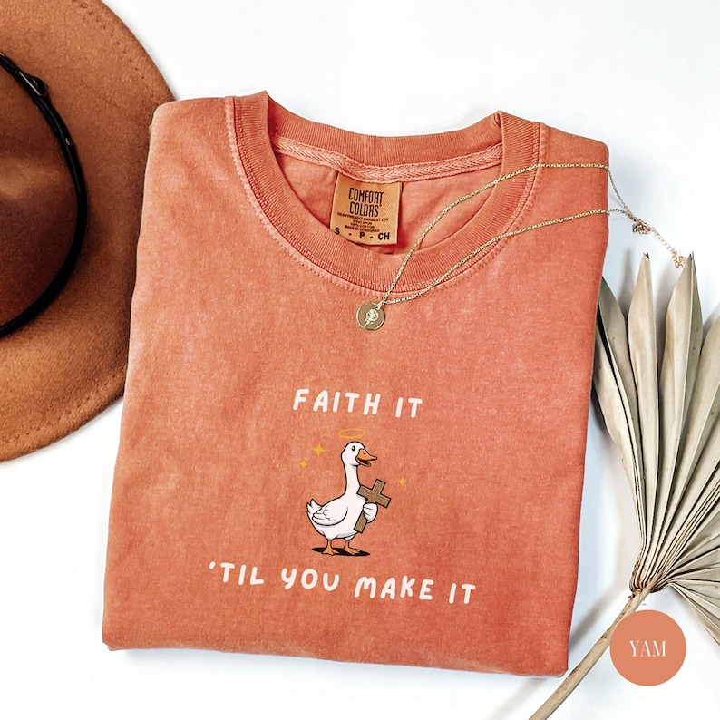 May include: A coral-colored Comfort Colors t-shirt with the phrase "Faith It 'Til You Make It" and a cartoon goose holding a cross. A gold necklace and a brown hat are also visible.