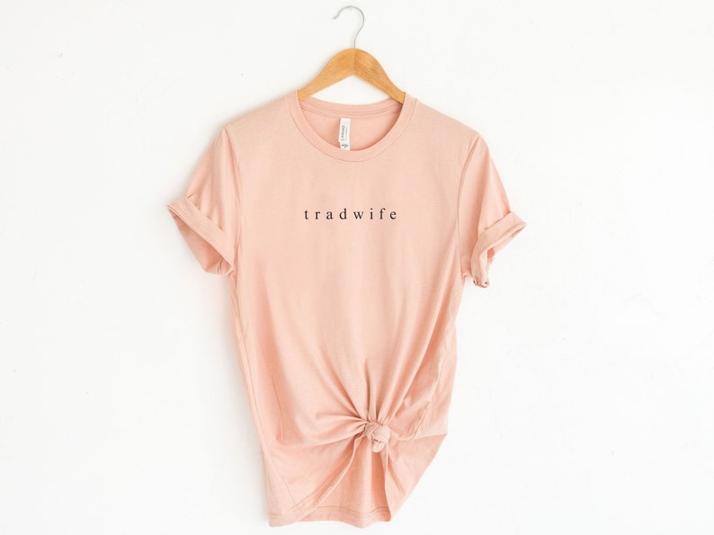 Op de afbeelding: Een lichtroze T-shirt met het woord "tradwife" in zwart op de voorkant.