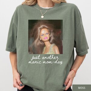 May include: Moss green oversized t-shirt with a square graphic of a doll with a distressed expression. The graphic includes the text "just another manic mom-day" in a handwritten style font.