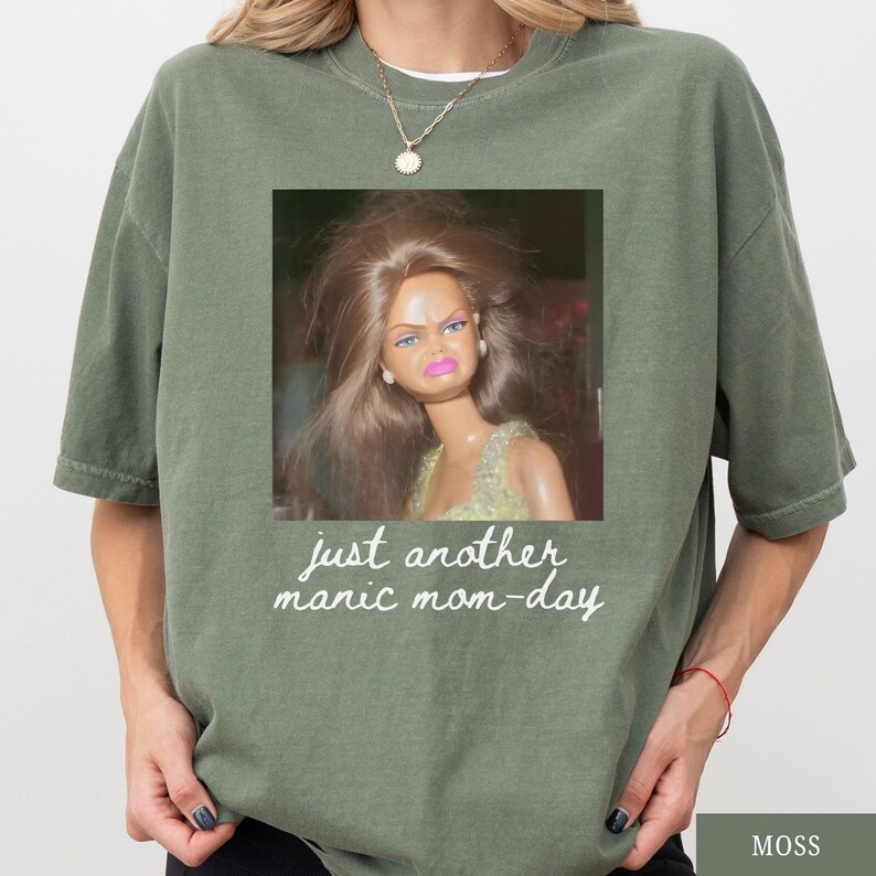 May include: Moss green oversized t-shirt with a square graphic of a doll with a distressed expression. The graphic includes the text "just another manic mom-day" in a handwritten style font.