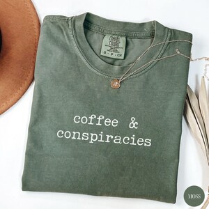 K&ouml;nnte beinhalten: Ein moosgr&uuml;nes T-Shirt mit dem wei&szlig;en Aufdruck "coffee & conspiracies". Eine goldene Halskette mit Anh&auml;nger liegt auf dem Shirt. Das Shirt ist gefaltet, und ein brauner Hut ist teilweise sichtbar.