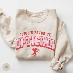 Puede incluir: Una sudadera beige con el texto "Cupid's Favorite Optician" en letras rosas y rojas. Un peque&ntilde;o cupido rojo con alas y una flecha est&aacute; debajo del texto.