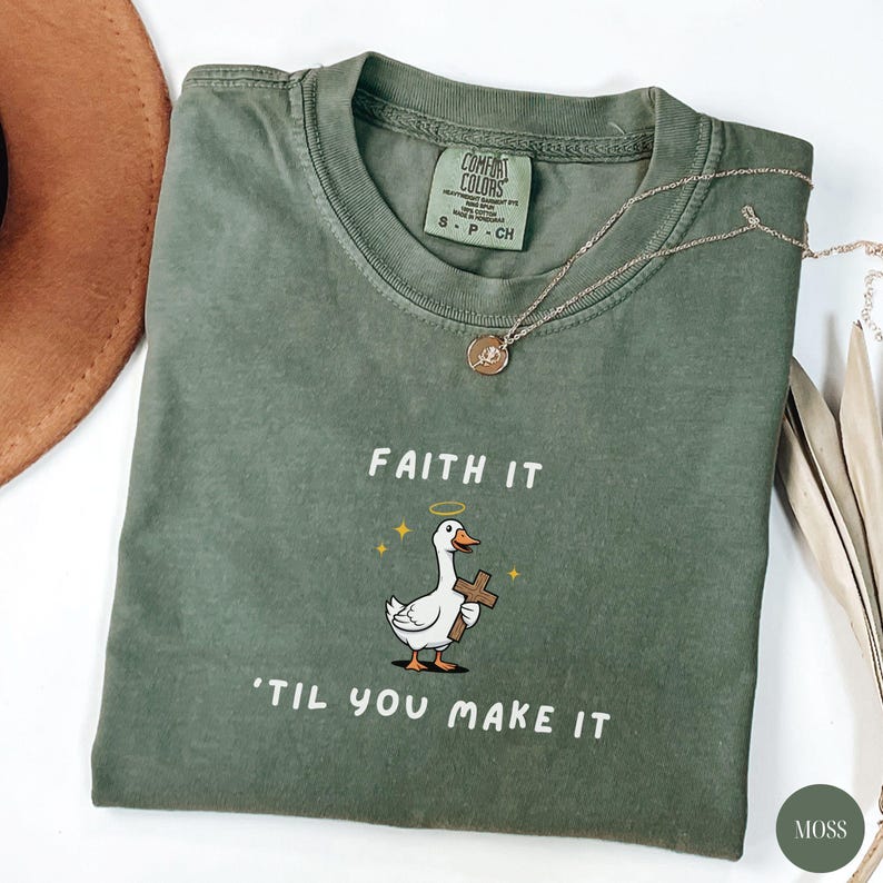 May include: A moss green t-shirt with the text "FAITH IT 'TIL YOU MAKE IT" and a cartoon goose holding a cross. The shirt has a crew neck and short sleeves. A gold necklace with a pendant is draped on the shirt.