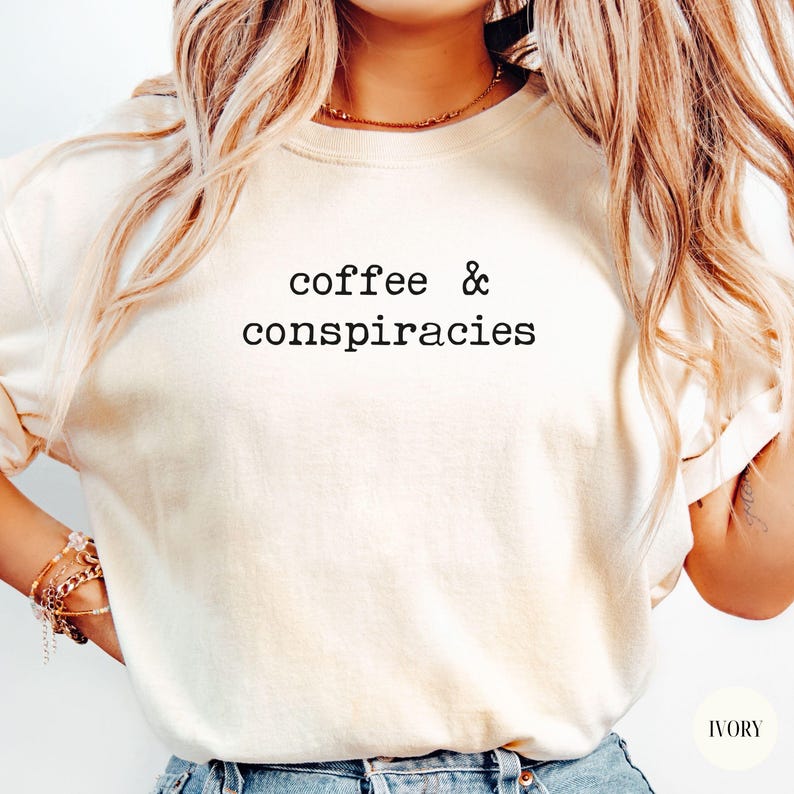 K&ouml;nnte beinhalten: Cremefarbenes T-Shirt mit Rundhalsausschnitt und dem schwarzen Text "coffee & conspiracies". Das Shirt hat eine neutrale Farbe und eine lockere Passform. Der Text ist mittig auf der Brust platziert. Es ist ein Freizeitkleidungsst&uuml;ck.