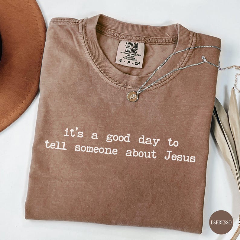 K&ouml;nnte beinhalten: Espressofarbenes T-Shirt mit dem wei&szlig;en Text "it's a good day to tell someone about Jesus". Das Shirt hat einen Rundhalsausschnitt und ein Comfort Colors-Etikett. Eine silberne Halskette mit Anh&auml;nger liegt auf dem Shirt.
