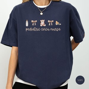 May include: A navy blue t-shirt with the text "pediatric cricu nurse" and a graphic of a baby bottle, two bows, a teddy bear, and a pacifier.