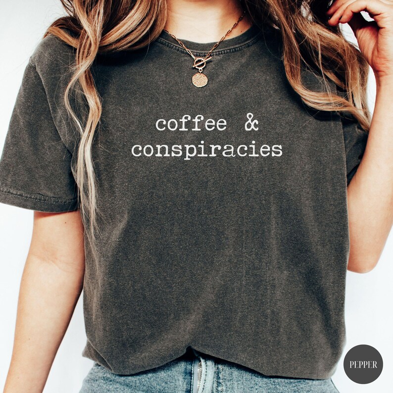 K&ouml;nnte beinhalten: Dunkelgraues T-Shirt mit dem wei&szlig;en Text "coffee & conspiracies". Das Shirt hat einen Rundhalsausschnitt und kurze &Auml;rmel. Das Model tr&auml;gt eine goldene Halskette. Das Shirt ist aus einem weichen, bequemen Material.