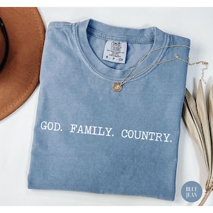 May include: A blue jean Comfort Colors t-shirt with the words "GOD. FAMILY. COUNTRY." in white. A gold necklace with a pendant rests on the shirt. The shirt is folded and has a small tag at the neckline.