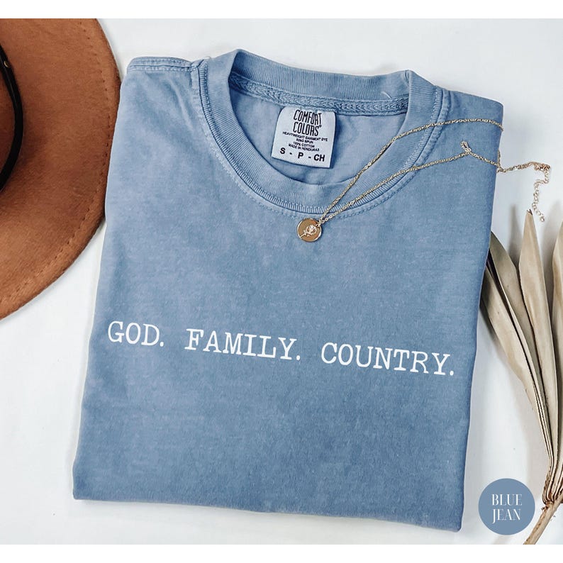 May include: A blue jean Comfort Colors t-shirt with the words "GOD. FAMILY. COUNTRY." in white. A gold necklace with a pendant rests on the shirt. The shirt is folded and has a small tag at the neckline.