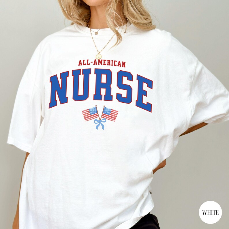 Op de afbeelding: Wit T-shirt met de tekst "ALL-AMERICAN NURSE" in rode en blauwe blokletters. Twee kleine Amerikaanse vlaggen met rode en blauwe strepen en een blauwe strik staan eronder. Het shirt heeft een casual stijl.