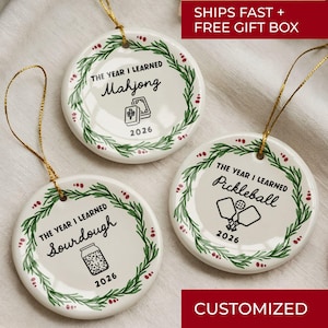 May include: Three round white ceramic ornaments with gold hanging strings. Each features a green wreath design, the text THE YEAR I LEARNED, the year 2026, and a graphic representing Mahjong, Sourdough, or Pickleball respectively.