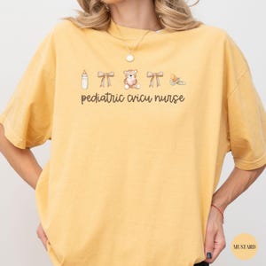 May include: A yellow t-shirt with the text "pediatric cricu nurse" and a graphic of a baby bottle, two bows, a teddy bear, and a pacifier.