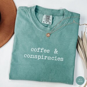 K&ouml;nnte beinhalten: Meergr&uuml;nes T-Shirt mit den wei&szlig;en Worten "coffee & conspiracies". Das Shirt hat einen Rundhalsausschnitt und kurze &Auml;rmel. Eine silberne Halskette mit rundem Anh&auml;nger liegt &uuml;ber dem Shirt. Das Shirt ist mit "Comfort Colors" gekennzeichnet.