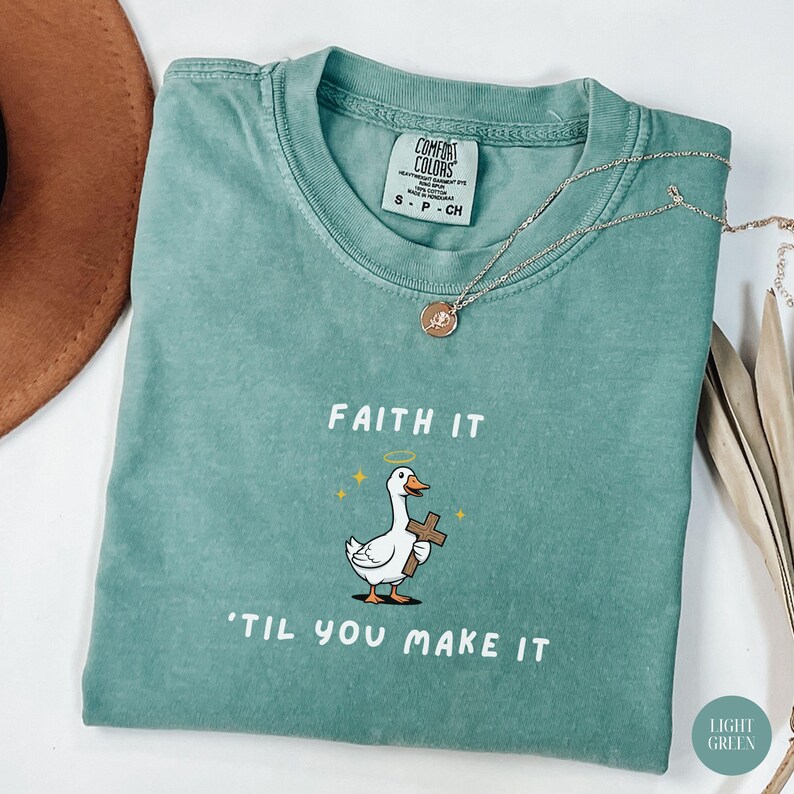 May include: Light green Comfort Colors t-shirt with the text "Faith It 'Til You Make It" and a cartoon goose holding a cross. A gold necklace with a pendant is draped across the shirt.