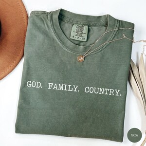 May include: A moss green Comfort Colors t-shirt with the words "GOD. FAMILY. COUNTRY." printed in white. A gold necklace with a pendant rests on the shirt. A brown hat is partially visible in the upper left corner.
