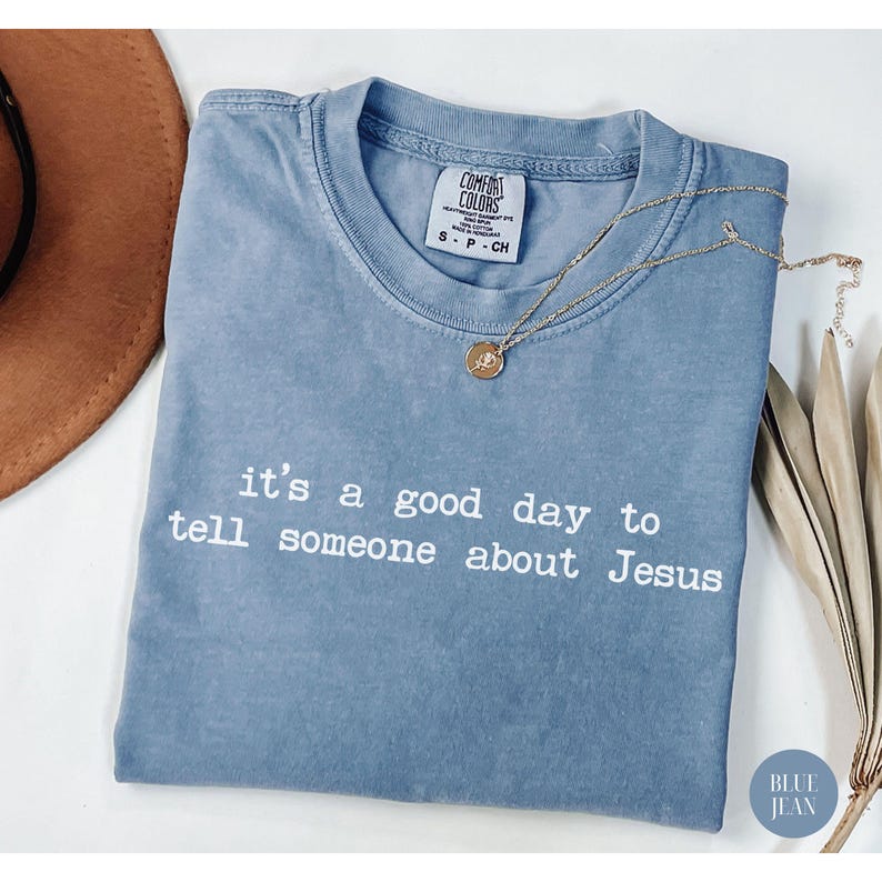 K&ouml;nnte beinhalten: Ein blaues Comfort Colors T-Shirt mit dem Text "it's a good day to tell someone about Jesus" in Wei&szlig;. Eine goldene Halskette mit Anh&auml;nger liegt auf dem Shirt. Das Shirt ist gefaltet und hat ein kleines Etikett.