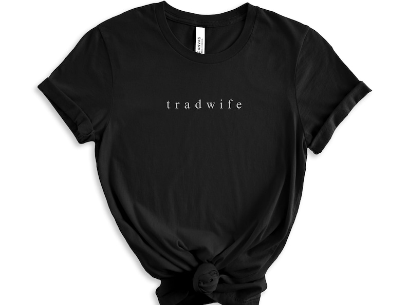 Op de afbeelding: Zwart T-shirt met de tekst "trad wife" in wit gedrukt op de voorkant.