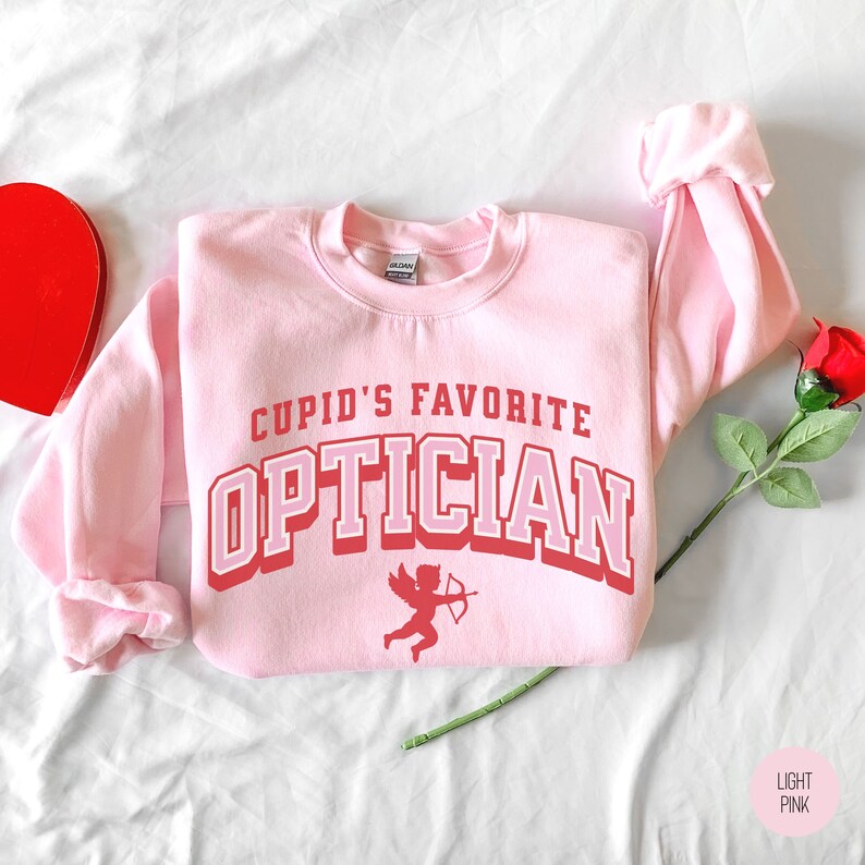 Puede incluir: Una sudadera rosa claro con el texto "Cupid's Favorite Optician" en letras rojas y blancas. Un peque&ntilde;o cupido rojo con una flecha est&aacute; debajo del texto.